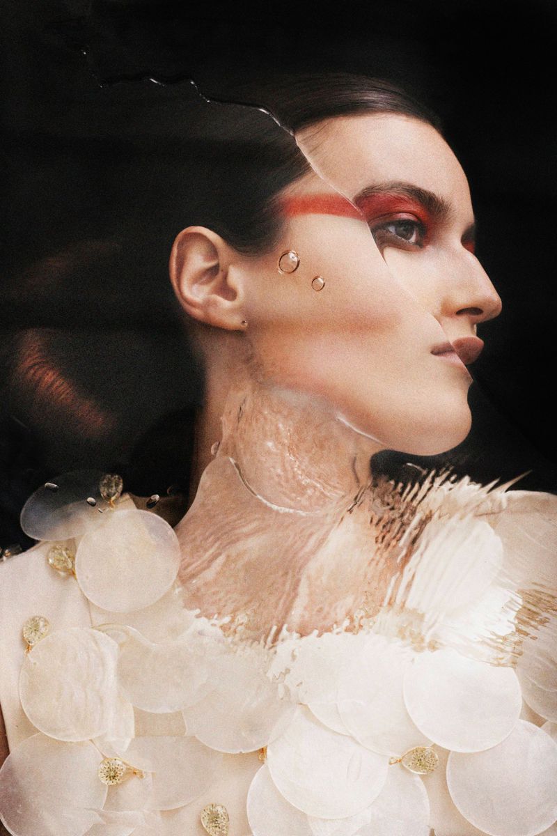 Model Noor Elliott in profile with red eye makeup and distorted glass refraction wearing a disc embellished top against black.