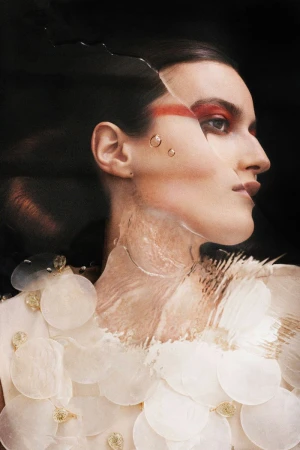 Model Noor Elliott in profile with red eye makeup and distorted glass refraction wearing a disc embellished top against black.
