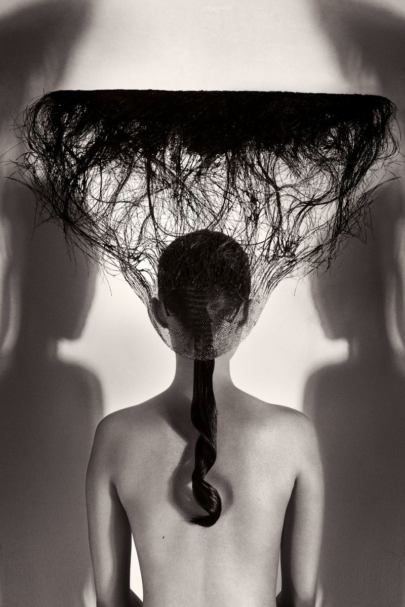 Back view of a topless figure with long dark hair and a horizontal line of hair overhead in a fine art photograph, black and white.