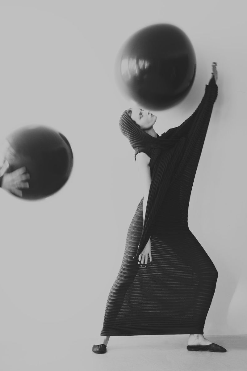Monochrome fashion shot: adult female in draped, hooded attire interacts with two floating black spheres against a plain backdrop.