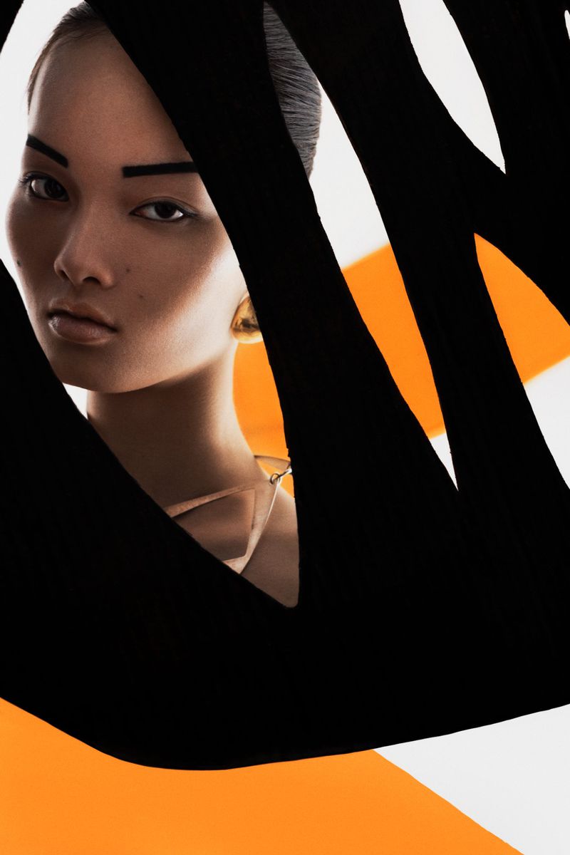 Portrait of an Asian woman with gold earrings posing in front of a white and orange background with black graphic design elements.