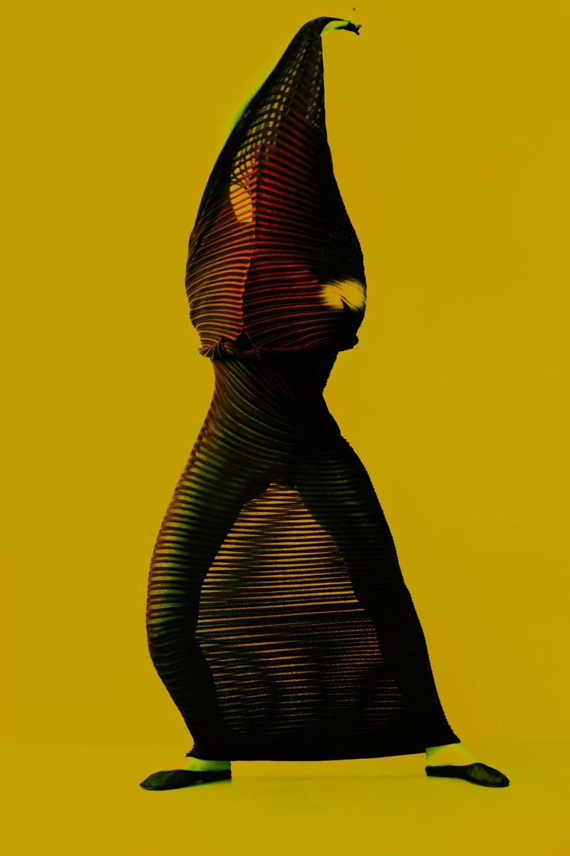 Person in a black pleated dress with an elongated hood-like head covering, striking a pose against a solid yellow backdrop.