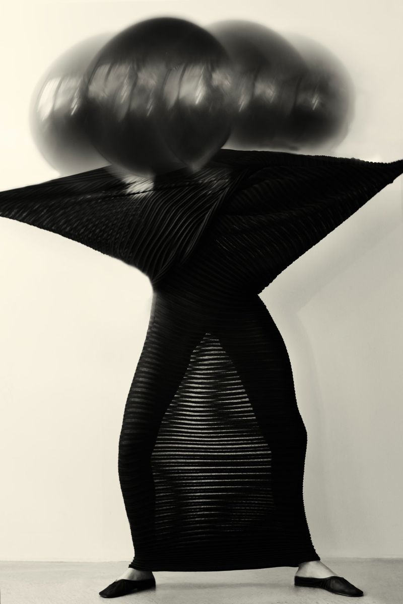 Woman in a flowing black dress poses with blurred black spheres above her head against a plain background in a studio setting.