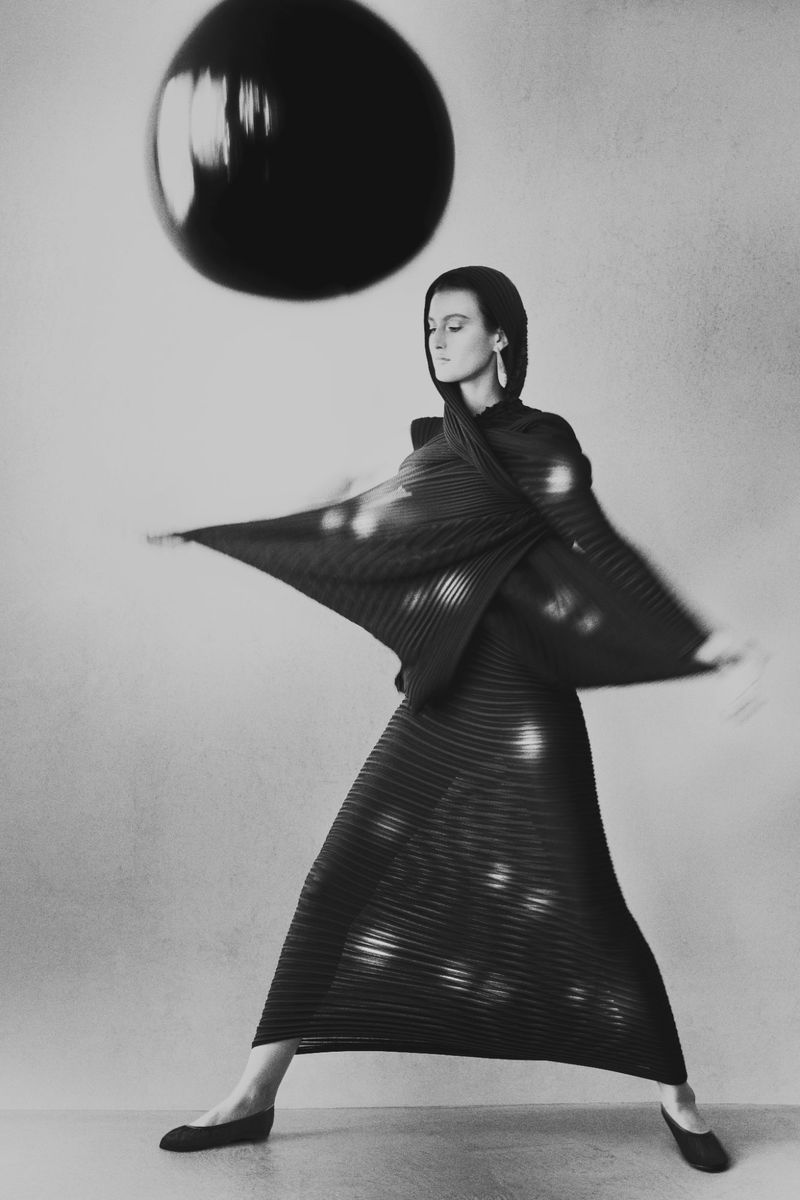 Fashion photography featuring a woman in a black pleated dress and head covering posing in front of a neutral backdrop with a large black balloon.