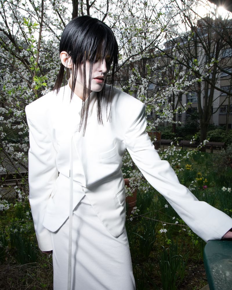 A person with dark hair and pale skin, wearing a white skirt suit, stands in an outdoor setting with trees and foliage in the background.