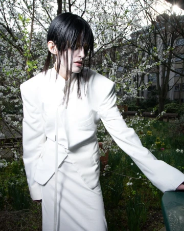 A person with dark hair and pale skin, wearing a white skirt suit, stands in an outdoor setting with trees and foliage in the background.