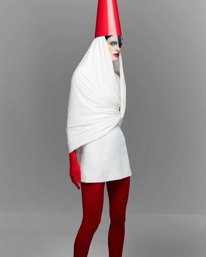 A fair-skinned person poses in a white mini dress with red tights and a red cone hat against a gray backdrop, fashion.