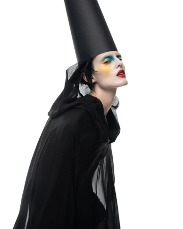 Elegant woman in black hooded cape and tall hat with vibrant makeup posing against a white backdrop in a fashion-forward artistic style.