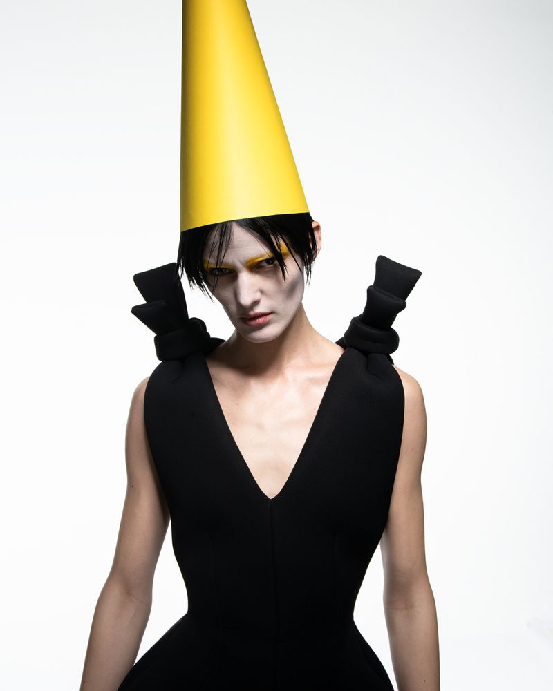 Person in black dress and yellow cone hat poses in a fashion studio setting, showcasing dramatic makeup and an eye-catching style.