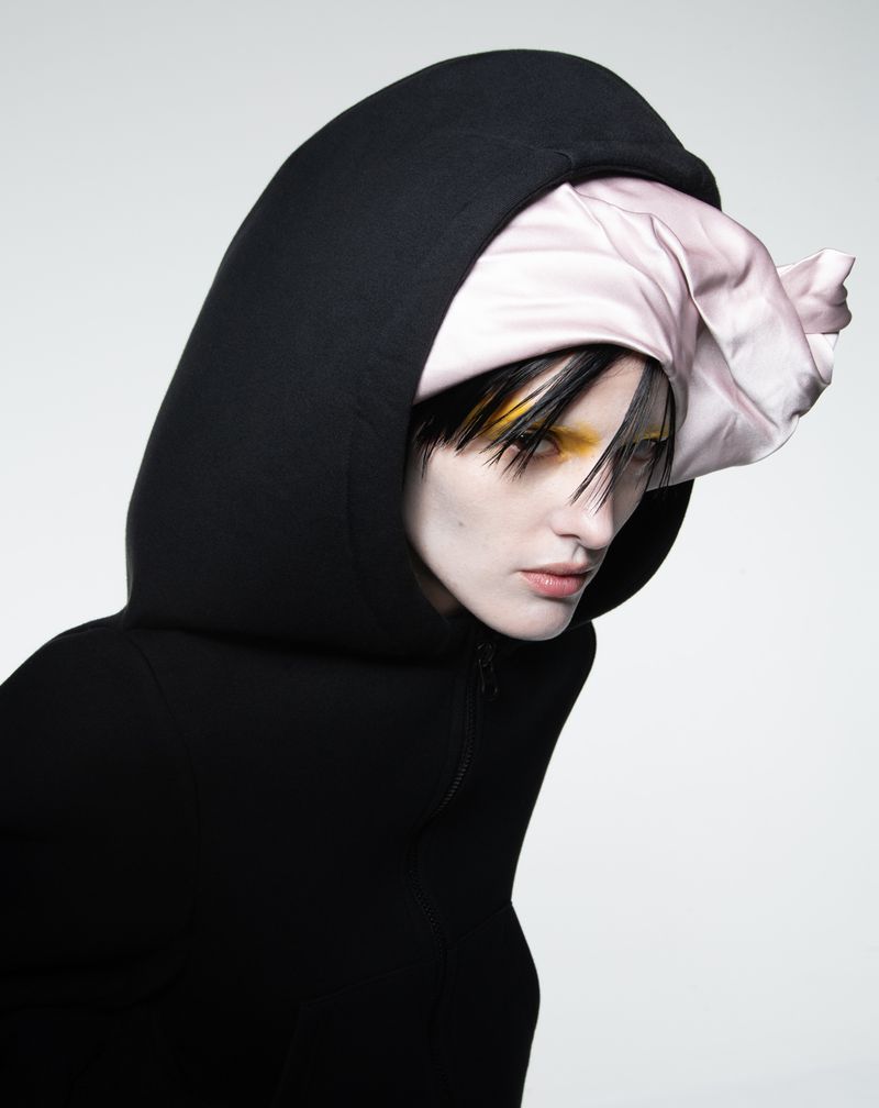 A person with yellow eye makeup and a silk head covering poses in a hooded black garment against a light gray backdrop in studio light.