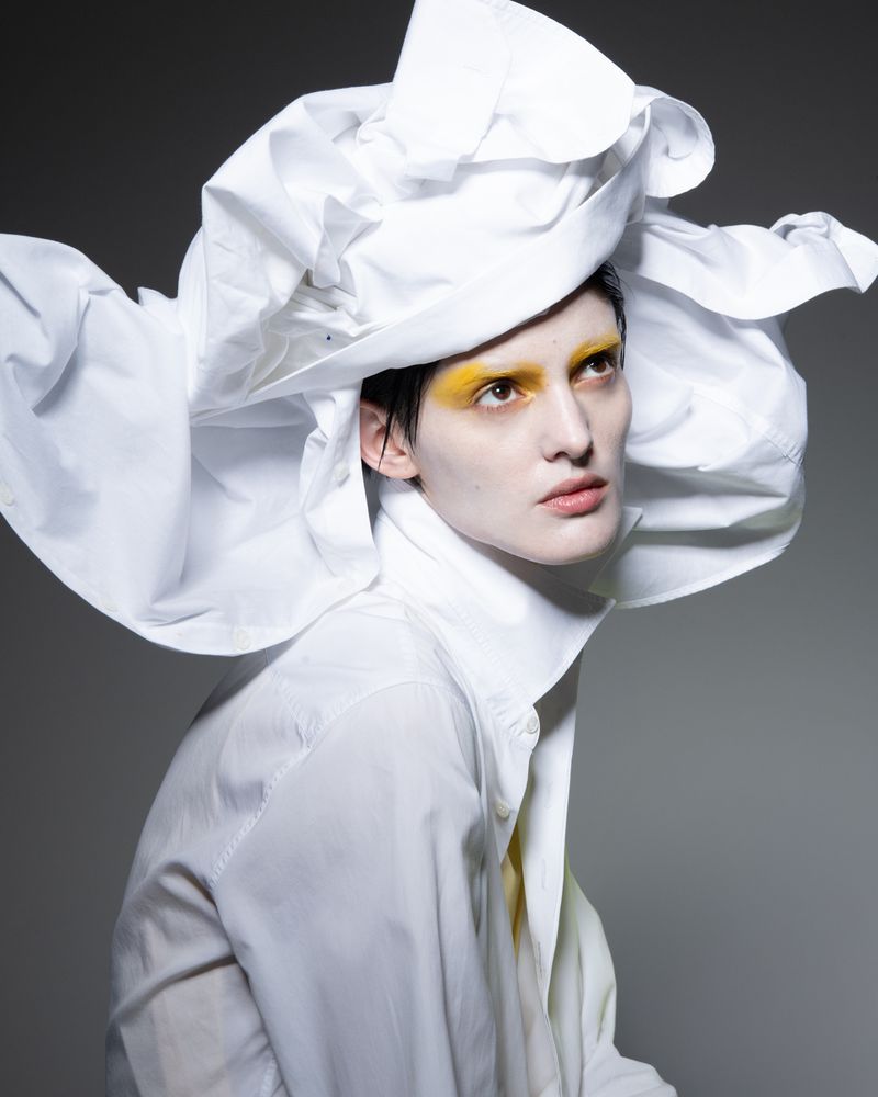 Fashion portrait of a person with white face paint, yellow eye makeup, and a large white ruffled hat and shirt against a grey background.