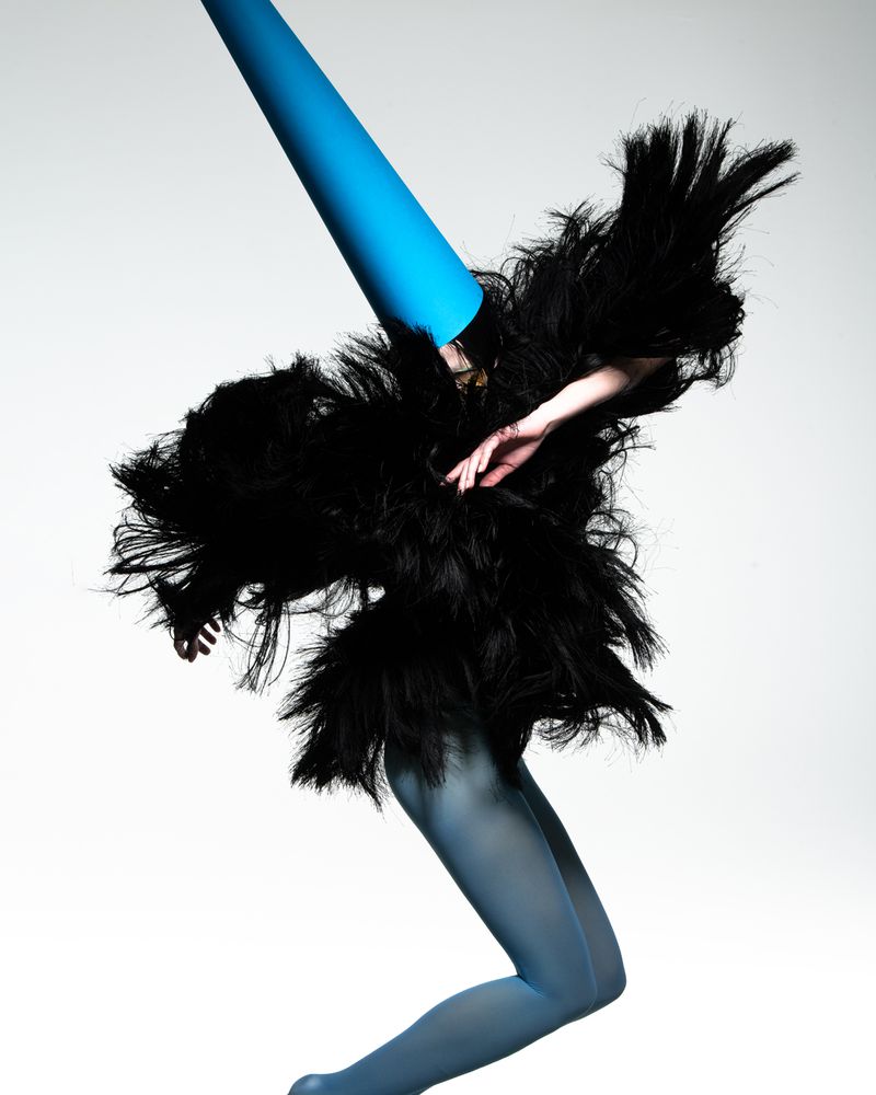Fashionable person with teal tights and a feathered black jacket poses in a studio setting. Head is covered by a blue paper cone.