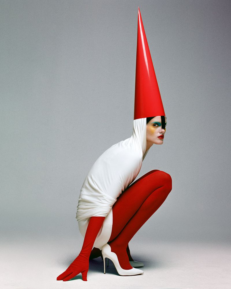 A woman in a white hooded dress, red tights, gloves, and a tall red hat poses in a studio with white heels and dramatic makeup.