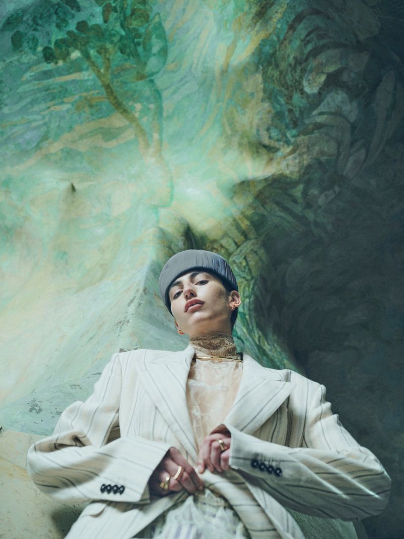 Low-angle portrait of a model in a cream pinstripe blazer and grey beanie against a painted fresco ceiling.