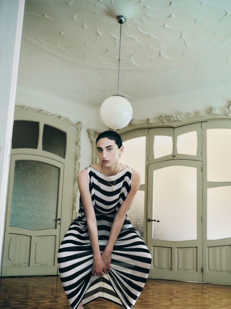 Fashion model in a black and white striped dress crouching beneath a round pendant lamp in an ornate room with arched doors.