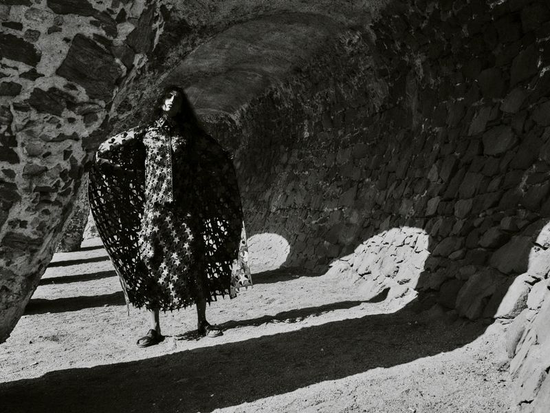 Model standing inside a stone archway wearing a dark patterned cape for Harper's Bazaar España.