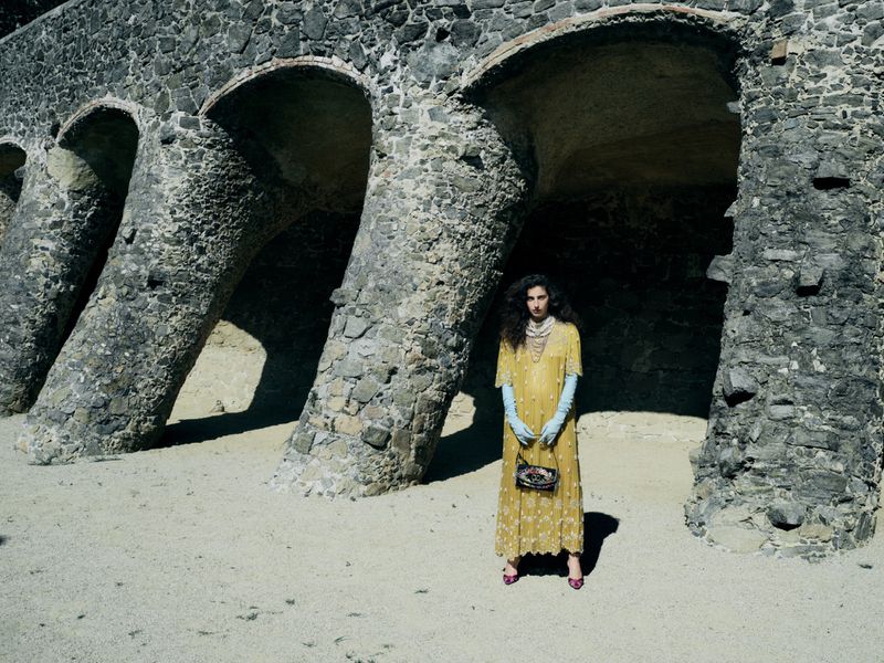 Woman in an embroidered yellow dress and light blue gloves standing beneath leaning stone arcade columns.