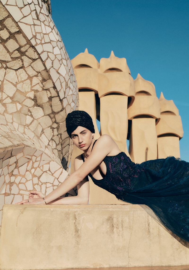Model wearing a black headwrap and dark embroidered gown reclining along a stone rooftop ledge before sculpted architecture.