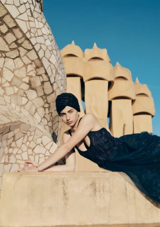 Model wearing a black headwrap and dark embroidered gown reclining along a stone rooftop ledge before sculpted architecture.