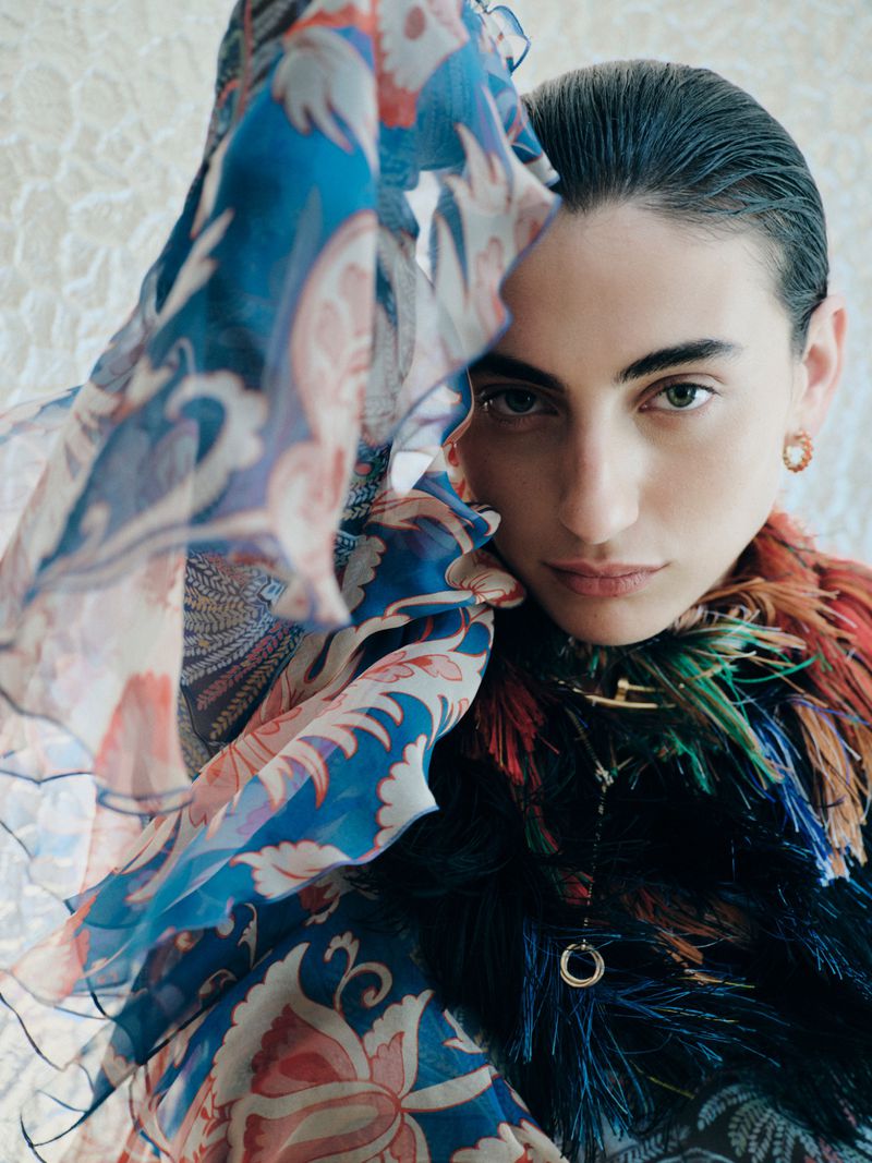 Model with slicked-back hair looking at camera framed by sheer blue printed chiffon and a multi-colored feather collar.