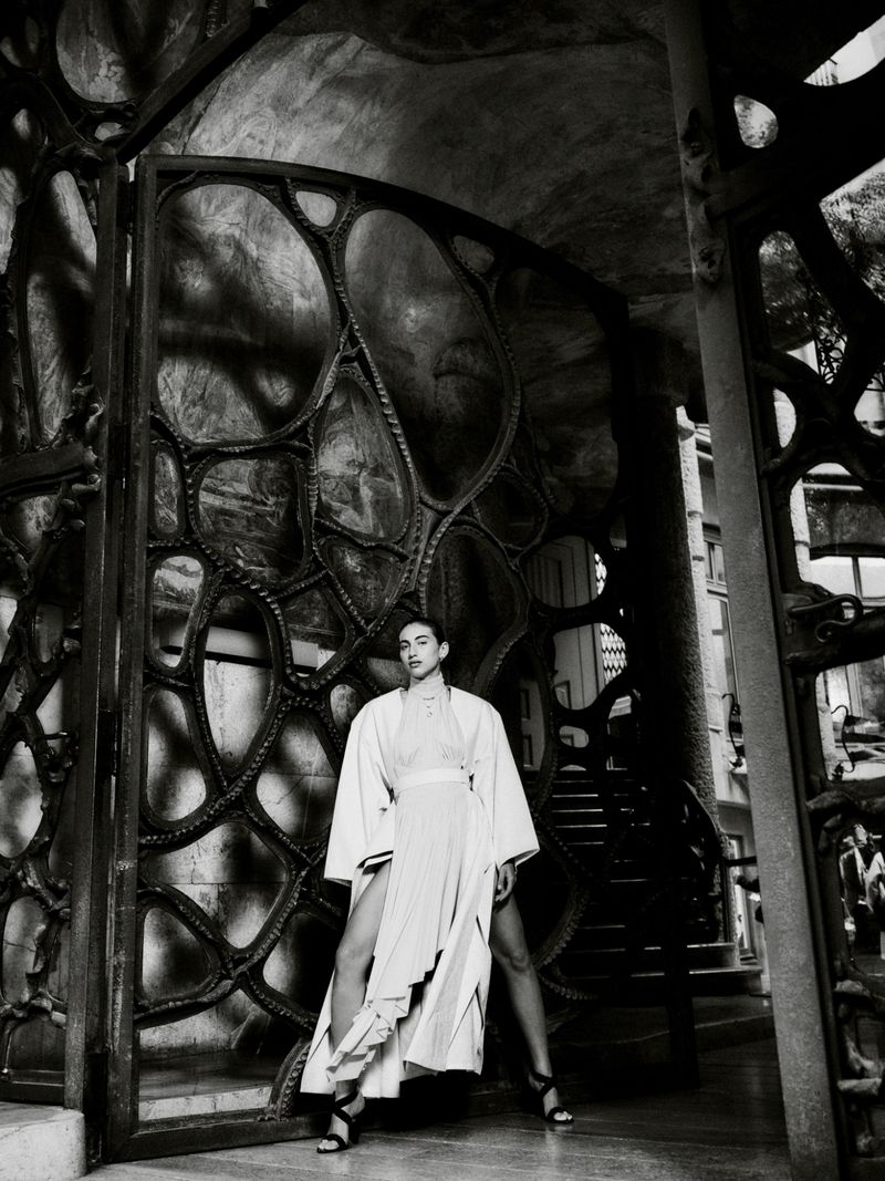 Model in a draped white cape coat and slit skirt posing inside an ornate Gaudí ironwork entranceway in black and white.