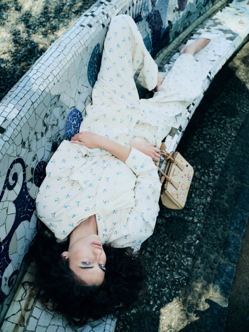 A woman with dark curly hair reclines on a blue and white mosaic bench, wearing a cream floral two-piece set with a beige handbag.