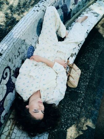 A woman with dark curly hair reclines on a blue and white mosaic bench, wearing a cream floral two-piece set with a beige handbag.