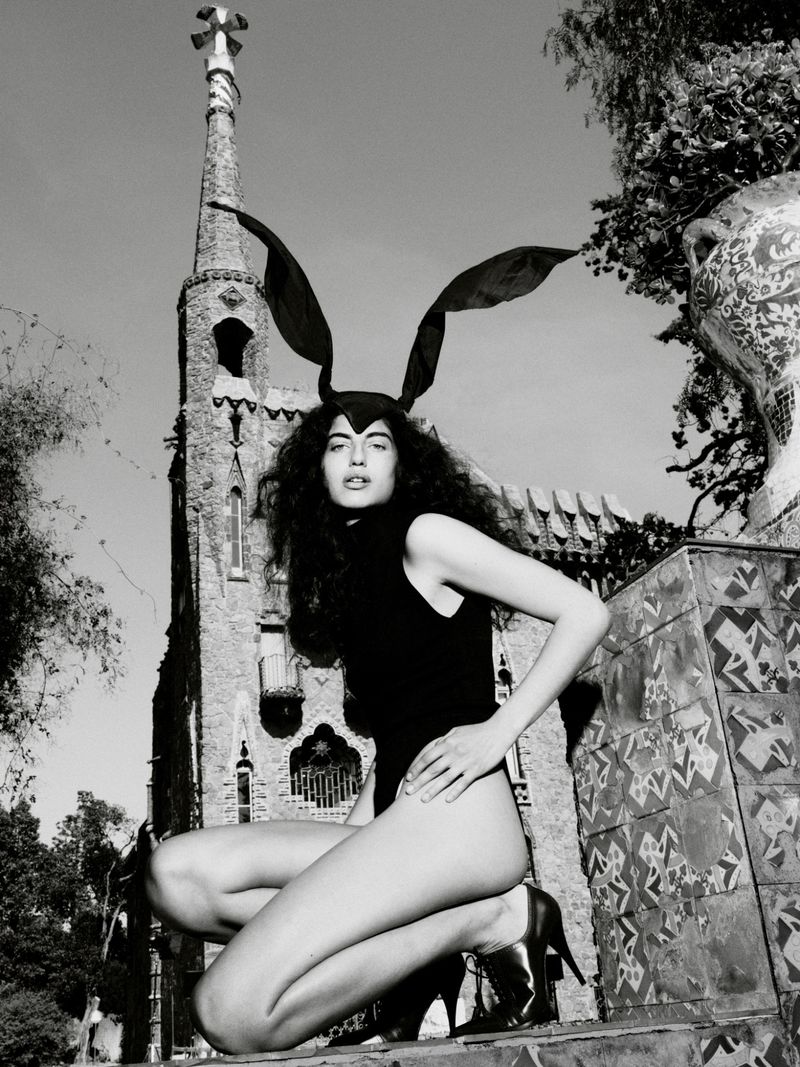 Model wearing dark bunny ears headband and black bodysuit crouching outdoors against a stone architectural tower in a black and white editorial photo.