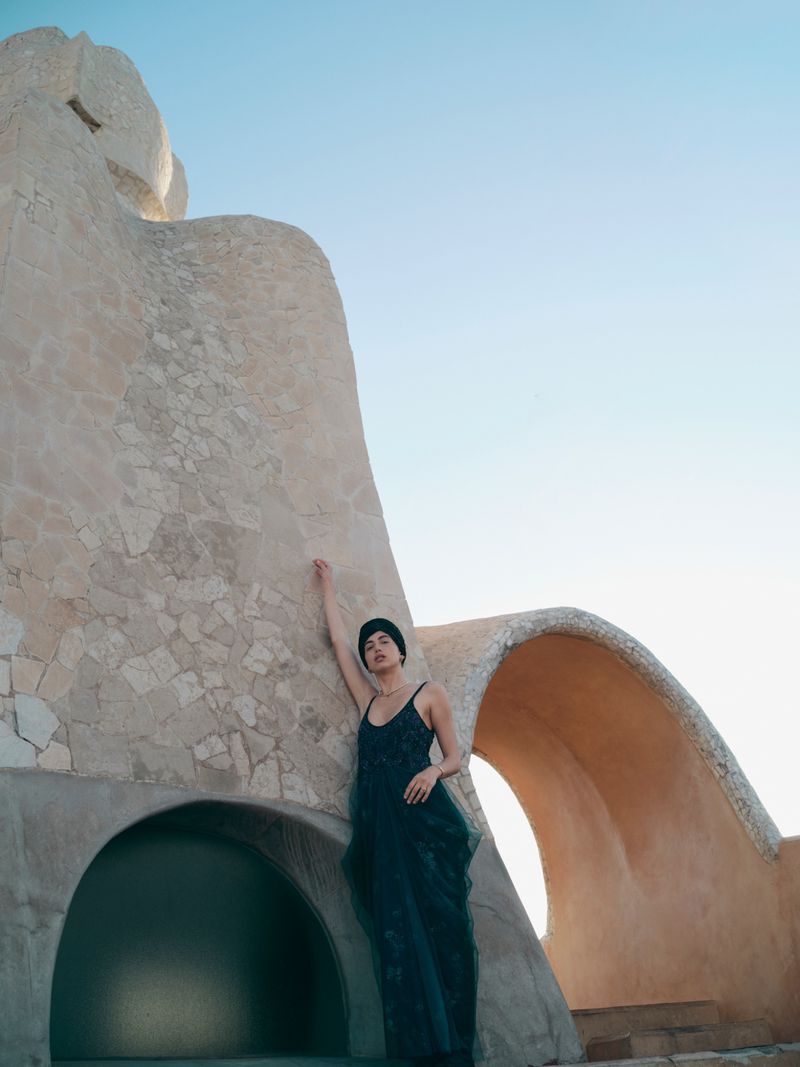 Model in a dark teal evening gown and black headwrap leaning against curved beige stone architecture under a clear sky.