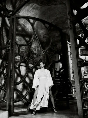 Model in a draped white cape coat and slit skirt posing inside an ornate Gaudí ironwork entranceway in black and white.