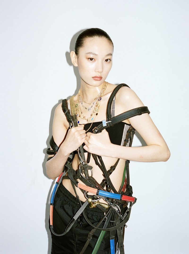 Woman in a black top and harness poses against a white background with natural lighting, showcasing fashion-forward, unique styling.