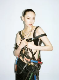 Model Fan Xu poses wearing layered leather equestrian harnesses and gold necklaces against a plain white studio backdrop.
