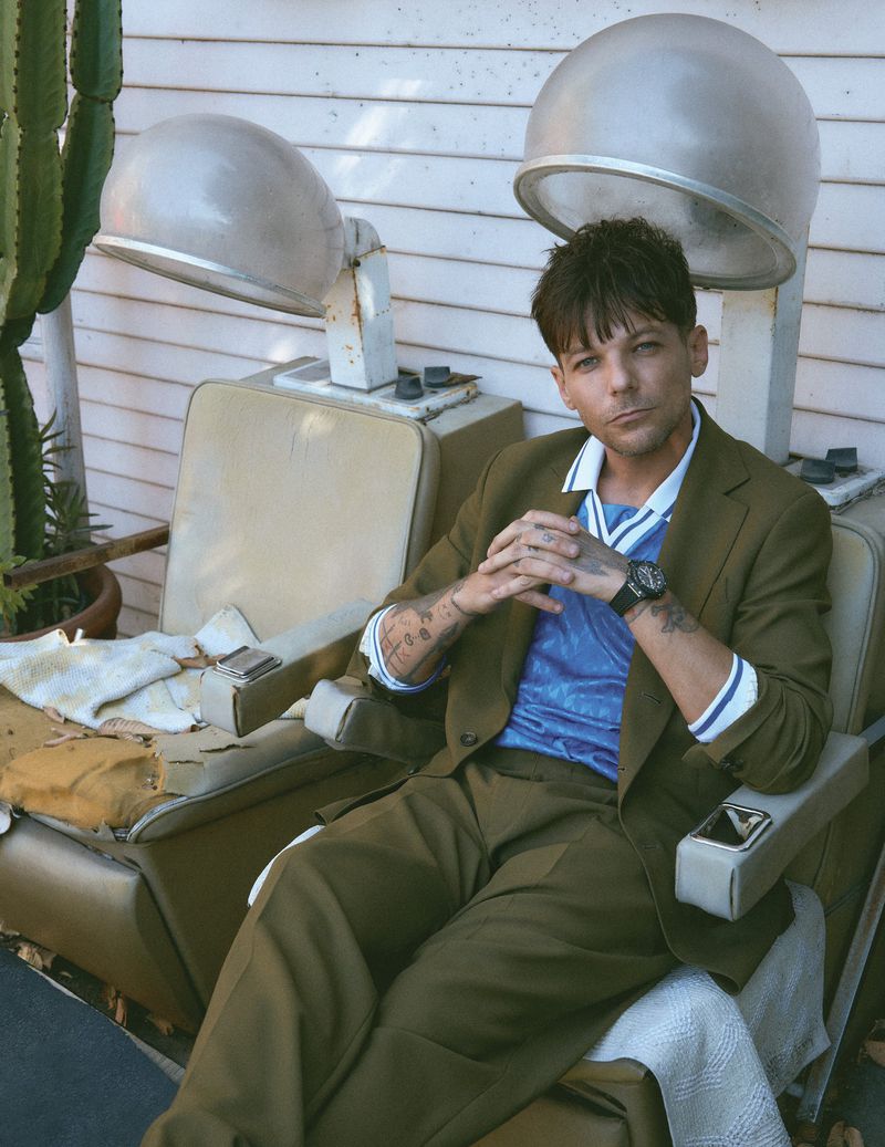 Louis Tomlinson seated in a vintage hair dryer chair wearing an olive green suit and blue polo shirt for Flaunt.