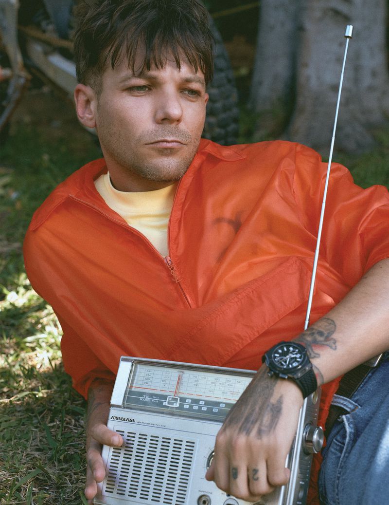Louis Tomlinson wearing an orange pullover lying on grass outdoors while holding a vintage portable radio with an extended antenna.