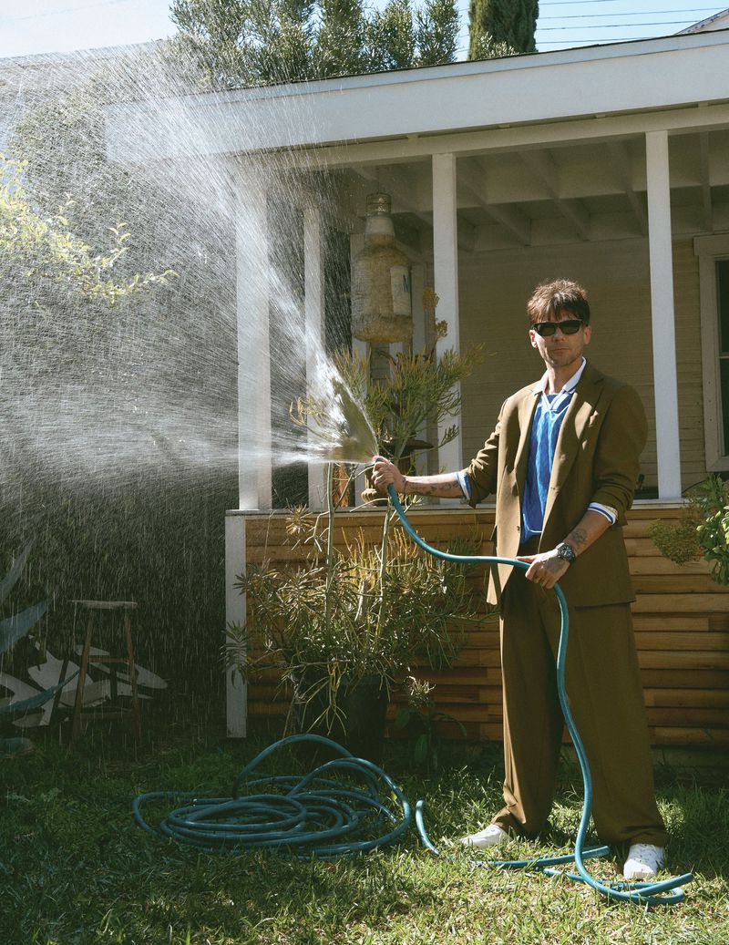 Louis Tomlinson in an olive suit and sunglasses spraying a garden hose in a front yard by Dean Isidro for Flaunt.