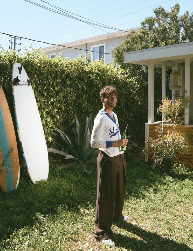 Louis Tomlinson standing on a sunny lawn wearing a white knit long-sleeve sweater and brown trousers next to surfboards.