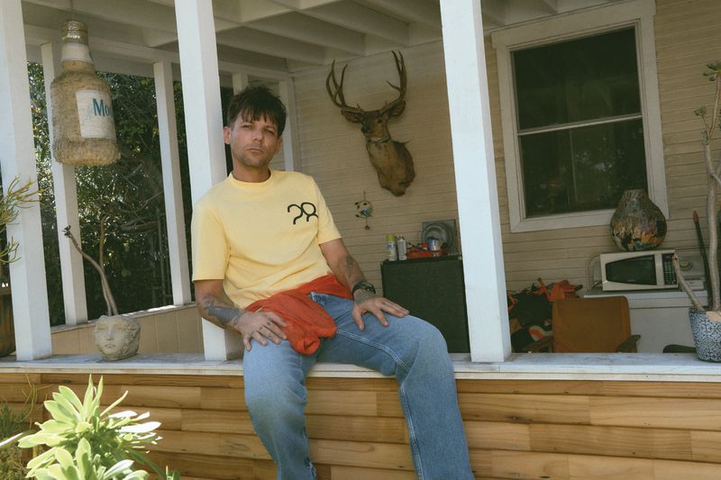 Louis Tomlinson sitting on a wooden porch ledge wearing a yellow t-shirt and blue jeans with a deer mount on the wall behind him.