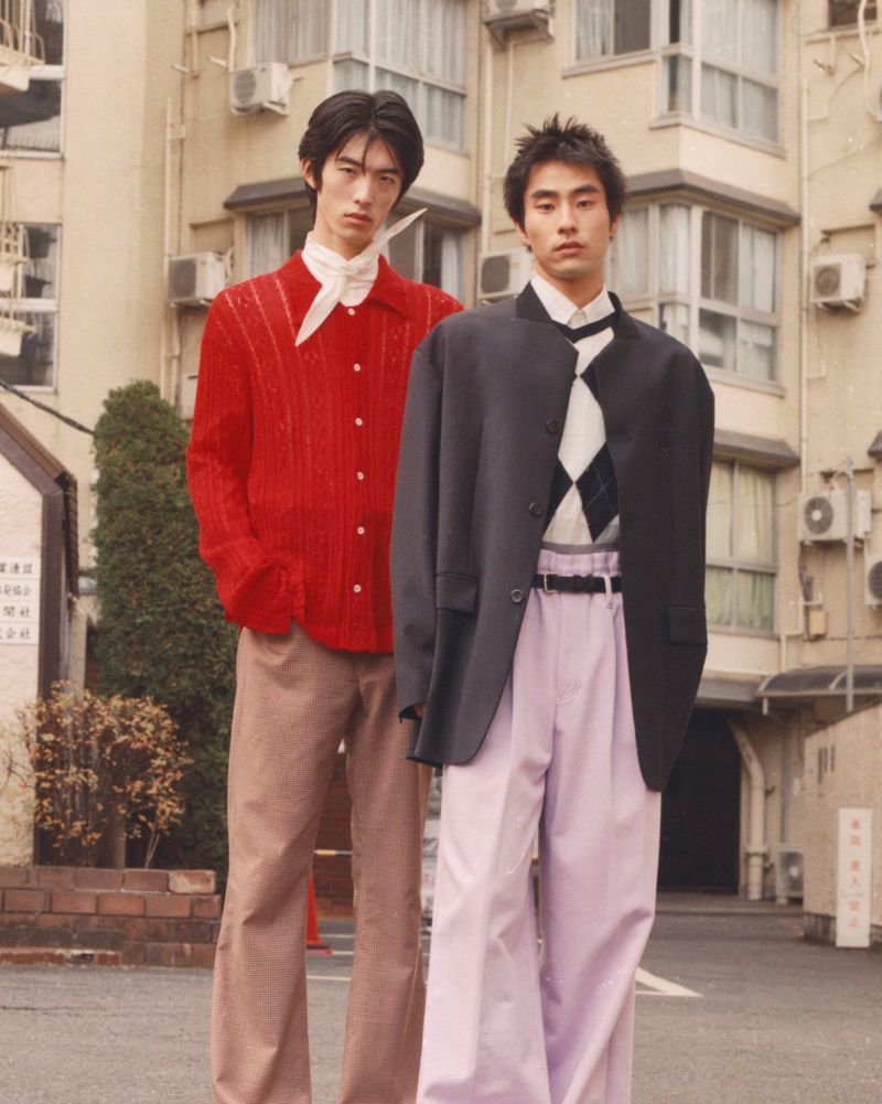Two young Asian men pose in fashionable outerwear against a muted urban backdrop, showcasing tailored garments and coordinated style.
