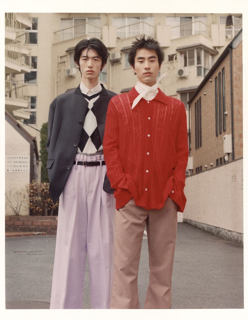 Two young Asian men in vintage fashion stand outdoors against a building backdrop, showcasing 70s-inspired shirts and wide-leg trousers.