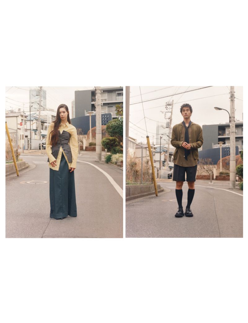 Two young adult models wearing casual fashion apparel stand on a residential street with buildings in the background.
