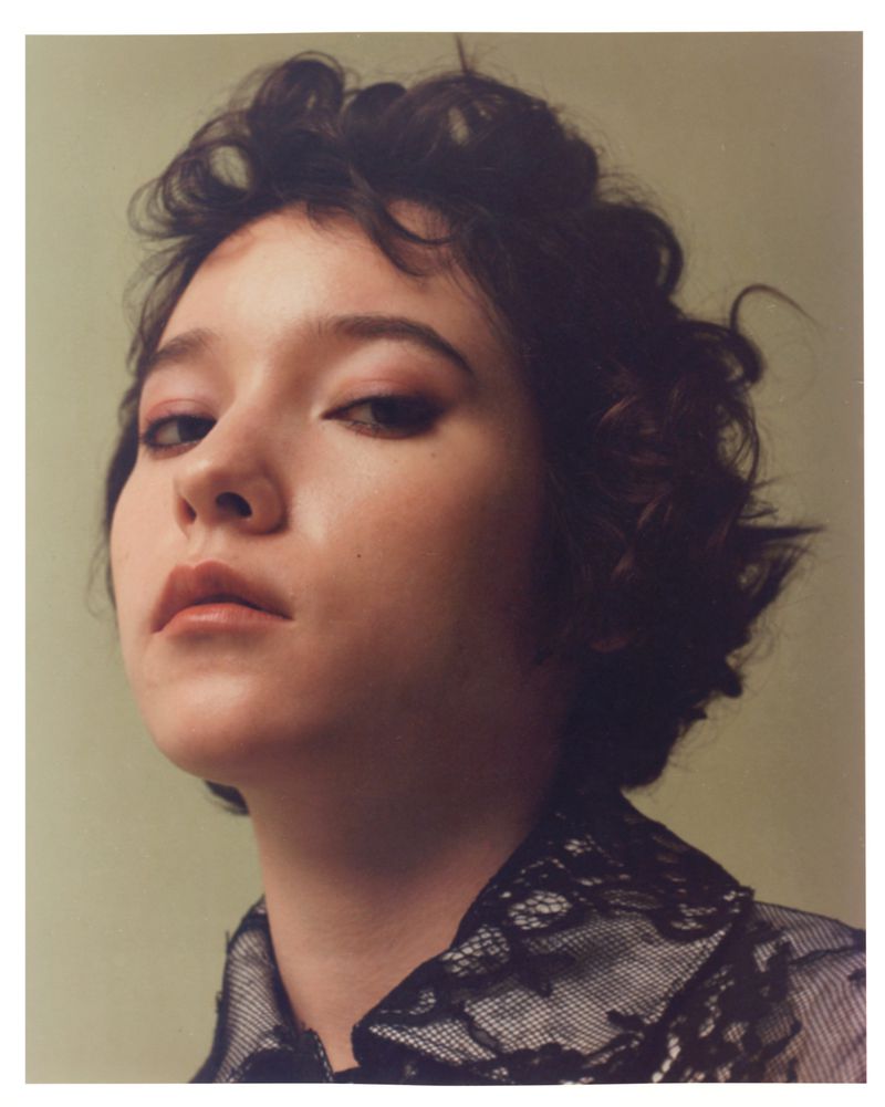 Portrait of a young woman with curly brown hair and fair skin, wearing a black lace top against a soft green backdrop in a studio setting.