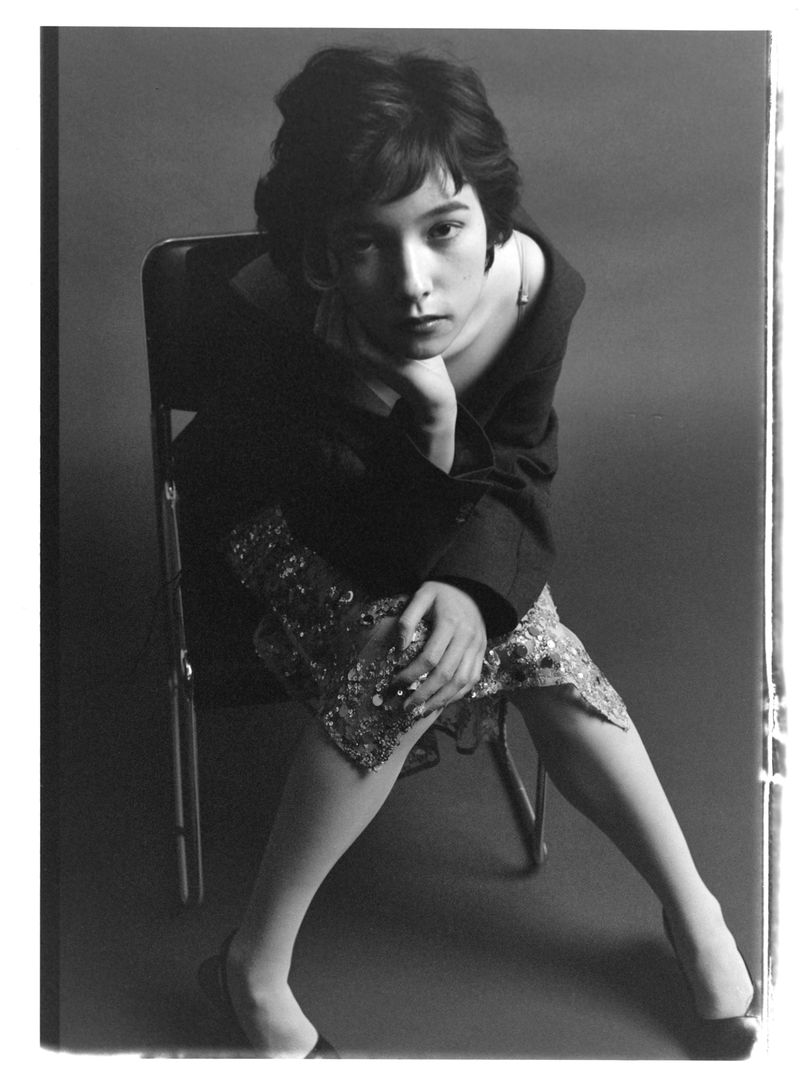 Black-and-white portrait of a young adult with short, dark hair sitting in a chair, gazing directly at the camera with a serious expression.