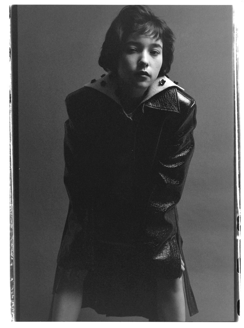 Monochrome portrait of a young adult female in a glossy black jacket, posed against a neutral grey background. Fashion shot.