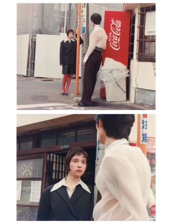 Two people in a street scene, one leaning against a post near a Coca-Cola vending machine, and the other in a black suit and red tights.