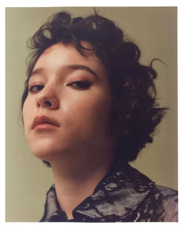 Close-up portrait of model Mikiya Nakano with short curly dark hair wearing a black lace shirt against a muted green backdrop.