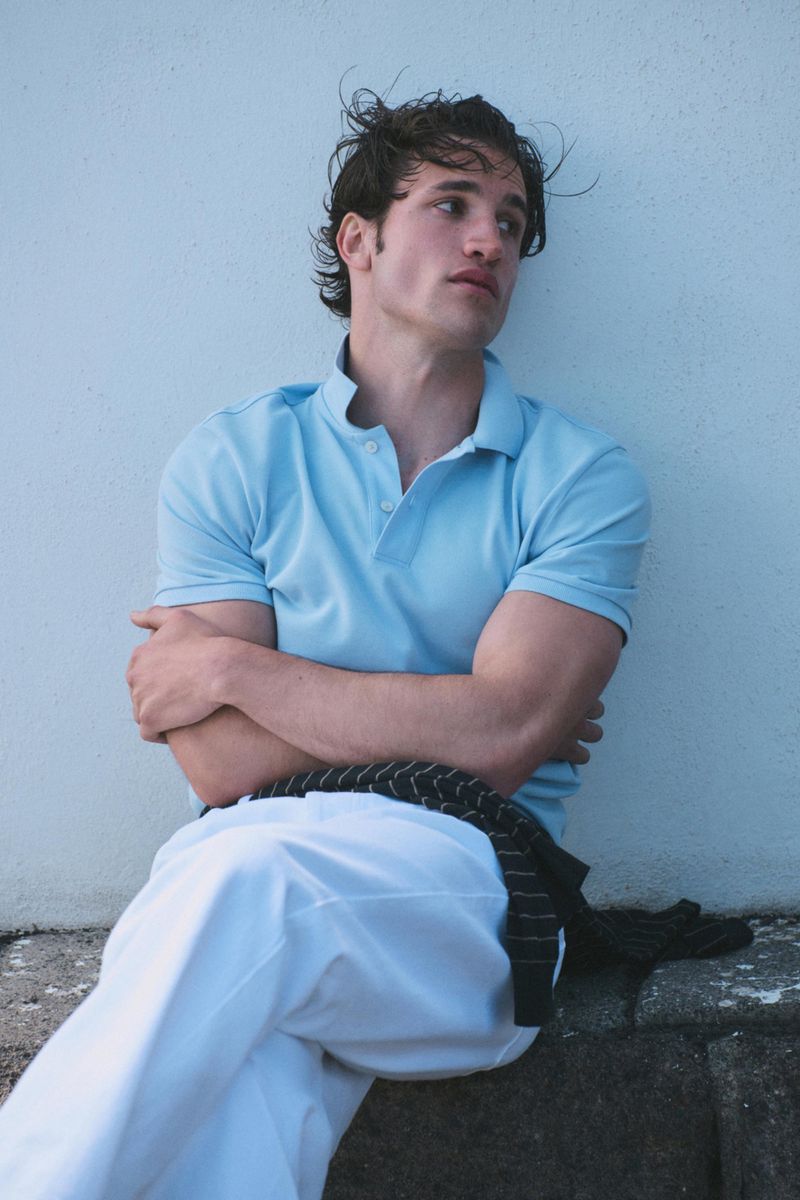 Model seated against a pale wall wearing a light blue polo shirt and white trousers with a striped sweater tied at his waist.