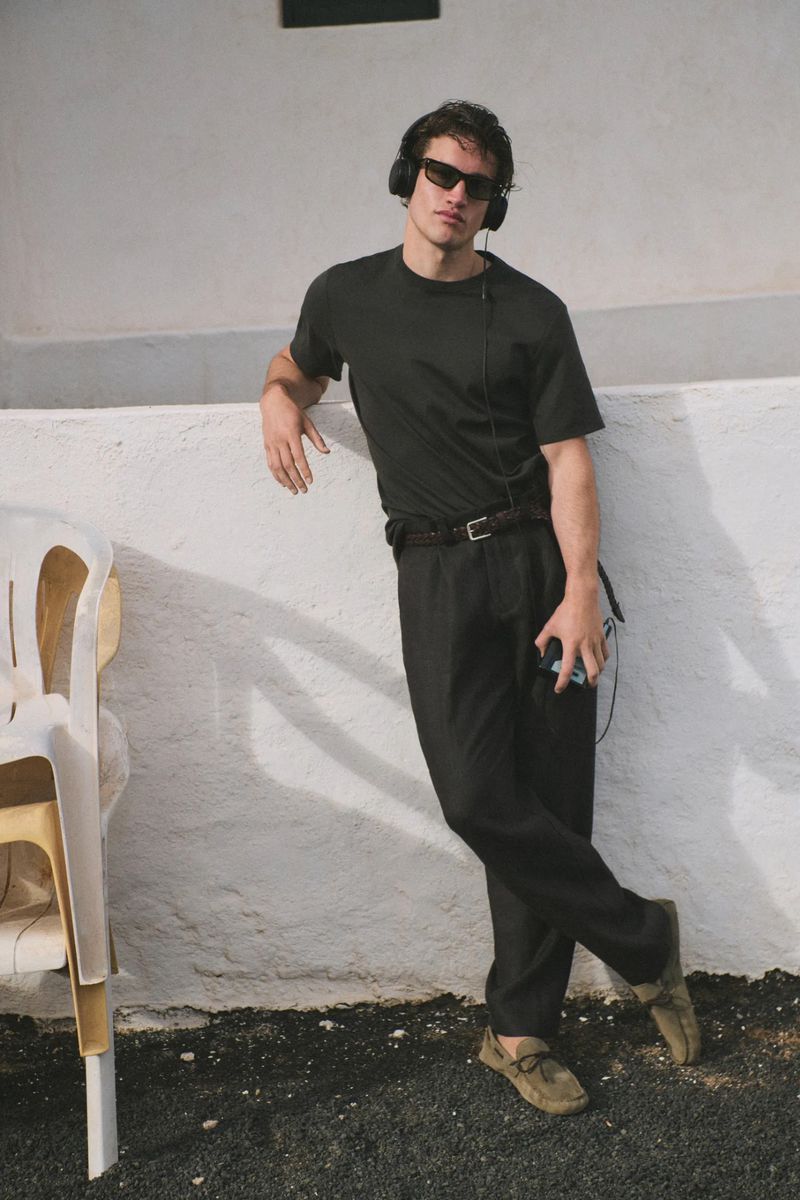 Man in black t-shirt, dark trousers, and sunglasses leaning against a whitewashed wall wearing over-ear headphones.