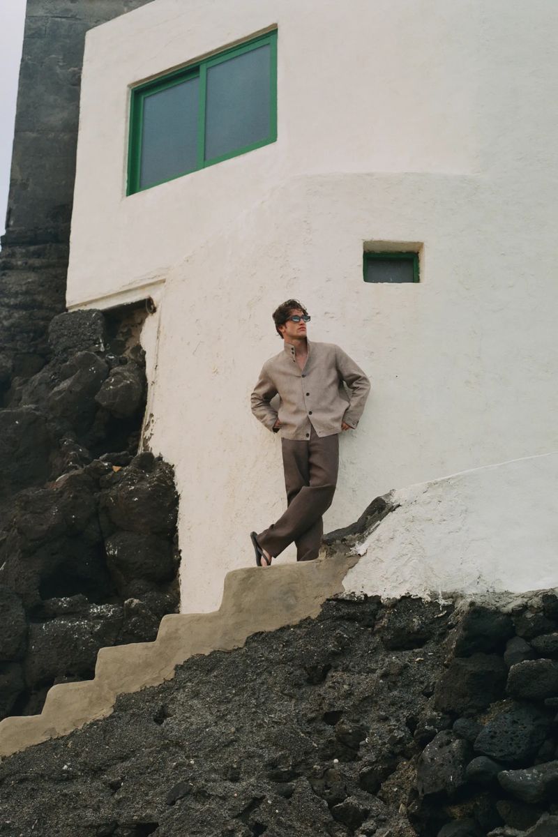 Model wearing a neutral beige buttoned jacket and brown trousers leaning against a white building wall on stone steps.