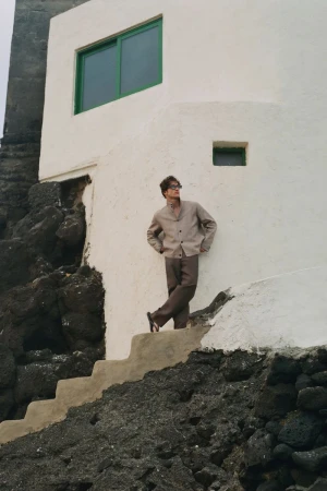 Model wearing a neutral beige buttoned jacket and brown trousers leaning against a white building wall on stone steps.