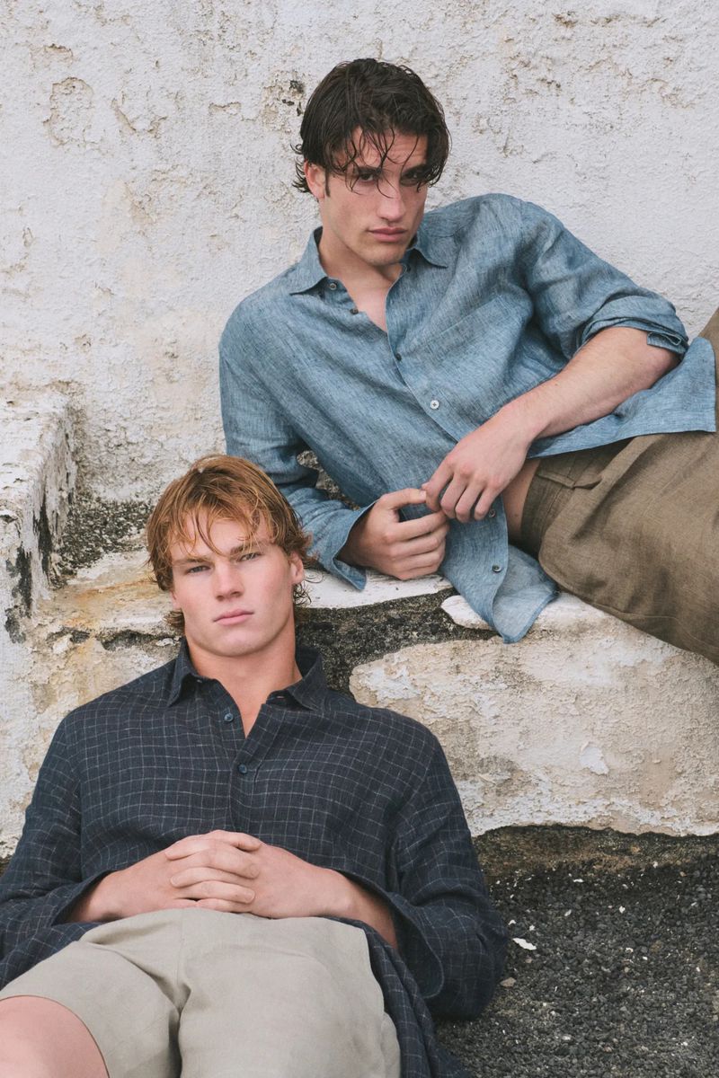 Two adult male fashion models pose in linen shirts and casual shorts on a stone staircase outdoors with sunlight. Editorial fashion.